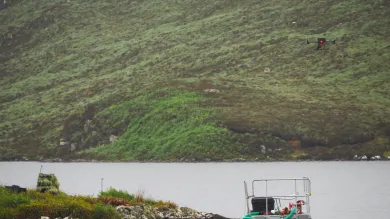 A drone hovers over a lake near a small floating platform with equipment, surrounded by lush green hills. The scene suggests a scientific or environmental study in a remote natural location.