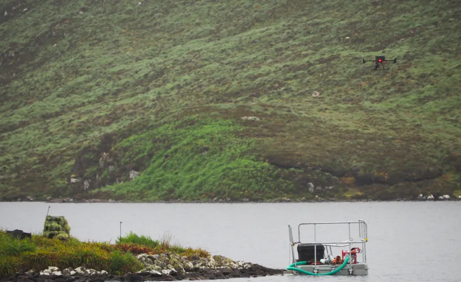 A drone hovers over a lake near a small floating platform with equipment, surrounded by lush green hills. The scene suggests a scientific or environmental study in a remote natural location.