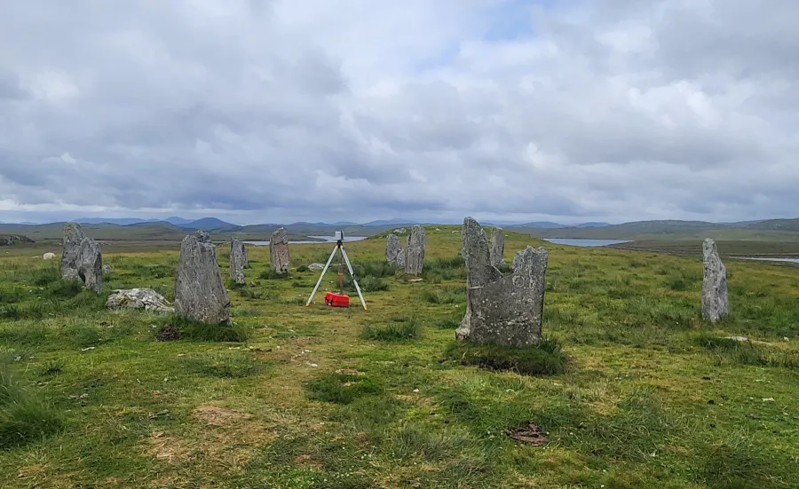 A grassy field features a circle of weathered standing stones, some covered in lichen. A tripod and red bag are positioned near the center under a cloudy sky.