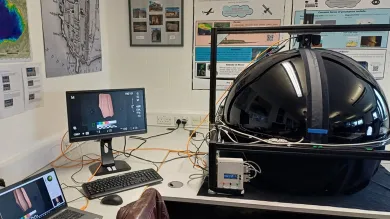 A laboratory setup includes a large black spherical device connected to wires and equipment, with two monitors showing images of an artifact. The room is decorated with posters and maps, suggesting a research or educational focus on archaeology.