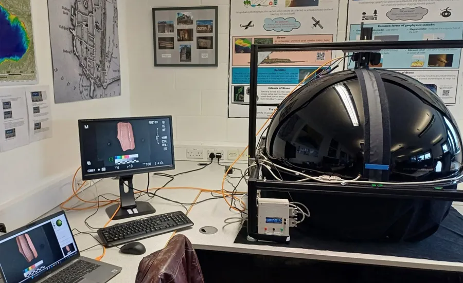 A laboratory setup includes a large black spherical device connected to wires and equipment, with two monitors showing images of an artifact. The room is decorated with posters and maps, suggesting a research or educational focus on archaeology.