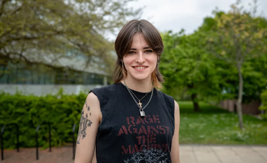 A student stands smiling outside the Physics building dressed casually in a sleeveless band T-shirt.