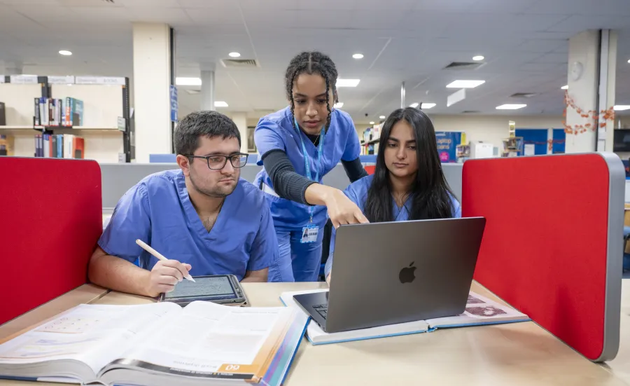 Medicine students wearing blue scrubs, study together in a library, with a laptop, tablet and reference book