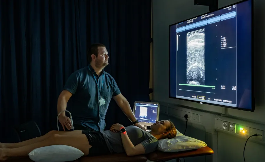 A medical professional uses an ultrasound device on a woman lying on an examination table, with a large screen displaying the ultrasound image.