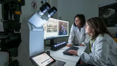 Two students in white lab coats using the microscopy lab facility. They're both analysing data on a computer monitor and there's a large microscope to the side of them.