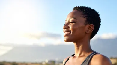 Person with short hair smiling with eyes closed against a bright sky background.