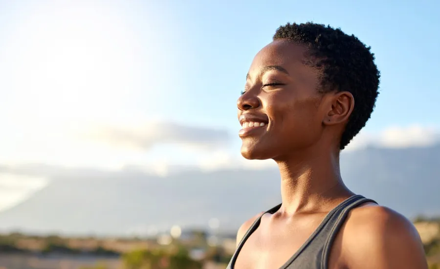Person with short hair smiling with eyes closed against a bright sky background.