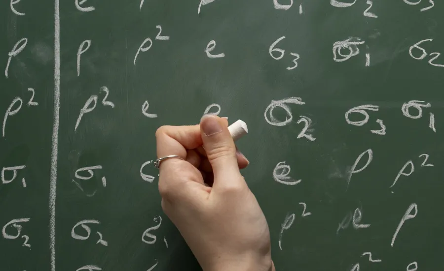 A blackboard showing digits written on in white chalk. A student stands in front of the board with her hand raised and holding the chalk.