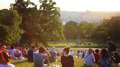 A large group of people sit on a grassy hillside in a park, facing a wide open lawn under warm, low sunlight. Trees frame the scene, and a distant cityscape is visible in the background.