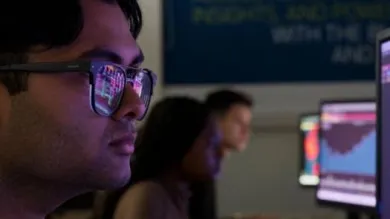 A student in the foreground looks intently at his PC screen, business analytics reflected in his glasses