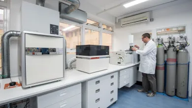 Laboratory technician in a white lab coat operating an Isoprime GeoVisION continuous flow isotope ratio mass spectrometer (IRMS) in a laboratory, with analytical instruments and workstations arranged along a bench.