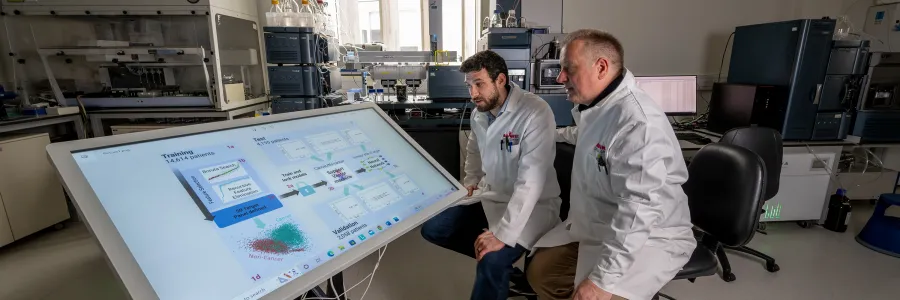 Two scientists sitting down looking at a large screen of graphs.