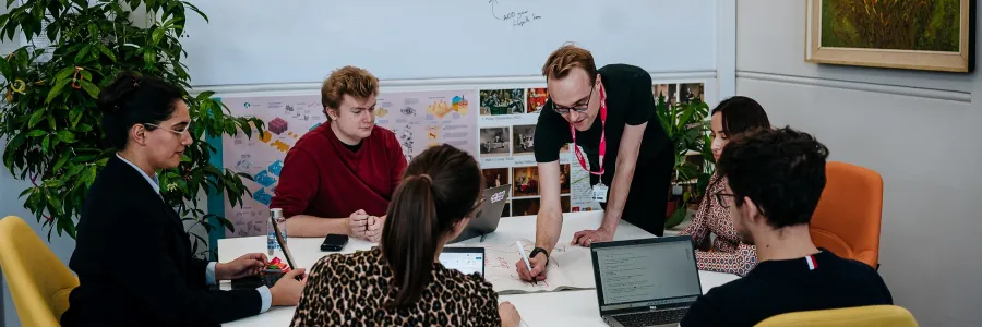 A group of people is collaborating around a table in a modern office space, with laptops, papers, and a whiteboard displaying diagrams and notes. One person is leaning over, writing in a book, while others focus on their discussions and screens.