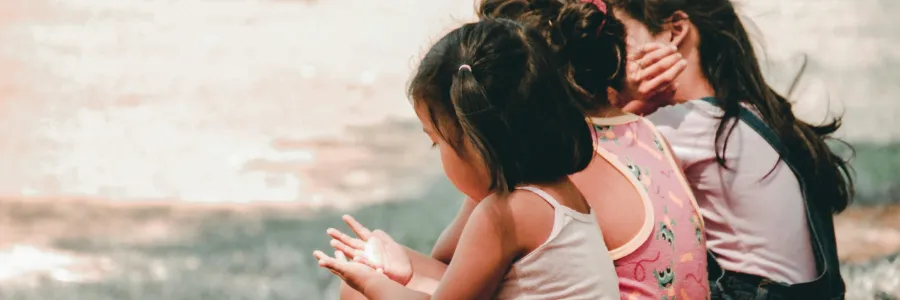 Three children sitting on grass, facing away, with colorful clothing.