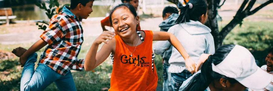 Children energetically playing outdoors, with a focus on a laughing girl in an orange shirt.