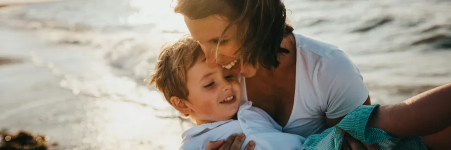 Woman holding a child on a sunny beach, both smiling.