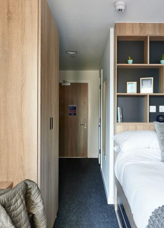 This image of a room at Southampton Crossings shows a large wardrobe against one wall, and a bed against the other. There is a desk in the corner and shelfing built above the headboard of the bed.