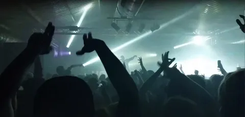 Silhouette of large crowd with their hands in the air, against bright lights of nightclub.