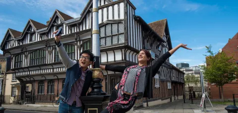 Two students take a selfie outside a large classic tudor house.