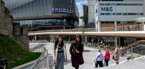 Two students talk whilst walking across a large paved area with several large shops.