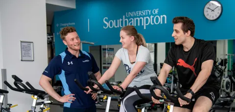 Two students working out on exercise bikes in a gym, assisted by a personal trainer.