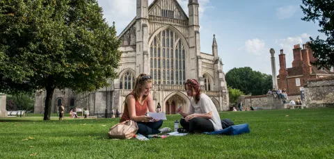 Two students sit talking on the grass outside a large, ornate cathedral.