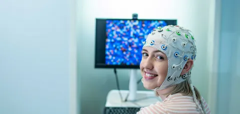 A student sits in front of a computer and turns to smile towards the camera. She is wearing a head cap that is connected to electrodes to monitor brain activity.