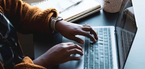 Fingers typing on a laptop with notepad, pen and mug in the background