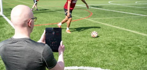 Dr Devon Lewis standing by a football pitch as a player kicks the ball. Devon is holding an iPad that shows live data streamed from a wearable sensor.