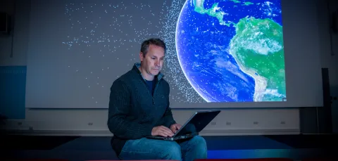 Professor Hugh Lewis sitting on the front bench of a lecture theatre as he consults his laptop. A graphic of satellites orbiting Earth is projected on the screen behind him.