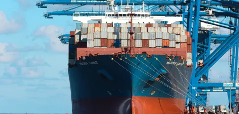 A heavily laden cargo ship sits under cranes by the dockside