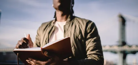A young Black man, whose face is unseen, writes in a notepad. He has a blue sky and a bridge that is out of focus behind him. The photo focuses on his hand with the pen.