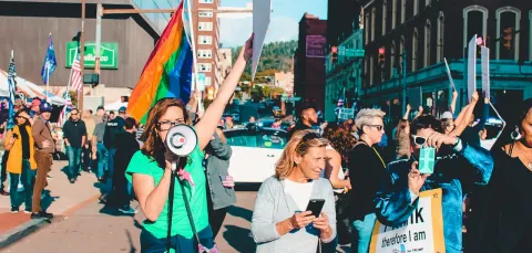 A diverse group of protestors, mainly women closest to us, hold up banners and a pride flag. One in the foreground is shouting into a loudspeaker.