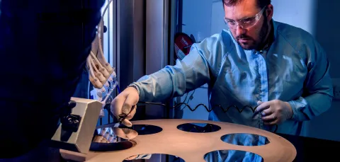 A bearded young scientist in protective overalls and glasses works with a flame hydrolysis deposition (FHD) system