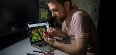 Bearded young researcher examines a circuit board while sitting at a desk