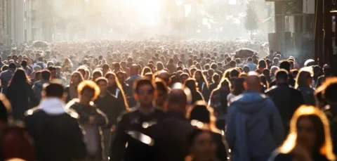 A large crowd of people stretches as far as the eye can see. A low, bright sun highlights a number of individuals who make up the crowd