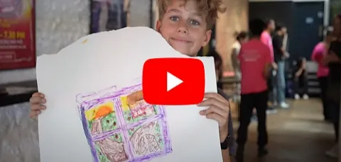 A boy holding a white banner with a drawing showing a view out of a window with the sun and green grass 