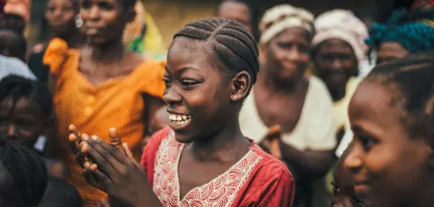 A young girl with braided hair claps and smiles amidst a group of villagers outdoors, capturing the vibrant spirit of Sierra Leone.