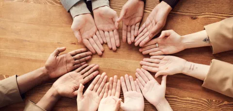 Several hands with varying skin tones turned palm-up in a circle on a wooden table, symbolizing diversity and unity.