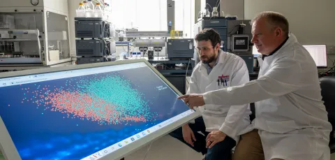 Two scientists observing blood test results on a screen. The results are colour coded in red and blue to show whether cancer is present or not.
