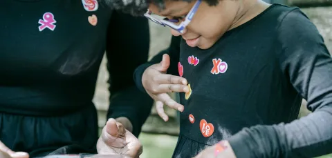 A young child is choosing stickers from a sheet held by an adult. The child is adding the colourful stickers to their top.  