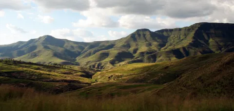 A landscape of rolling hills with long grass in the Lesotho Highlands. There is a mountain range on the horizon, and the sky is mainly cloudy with some clear blue areas.