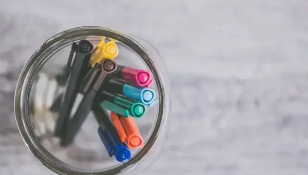 Top-down view of colourful pens in a glass jar on a grey wooden surface.