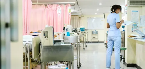 A hospital ward showing a trolley of equipment at the end of each bed. There is 1 member of staff facing away from the camera completing a chart.