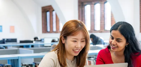 Two students sitting at a table in a library. They are smiling and looking at a laptop.