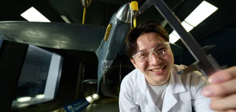 A smiling University of Southampton student in a lab coat and safety glasses, holding a caliper towards the camera. Behind them is a 1/6th scale Spitfire model that was put together for a 3rd year individual project in the University's R J Mitchell Wind Tunnel on Highfield Campus.