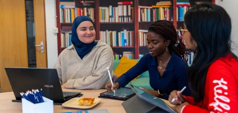 Nora and 2 friends study together in a library. They have their laptops open on the table in front of them.