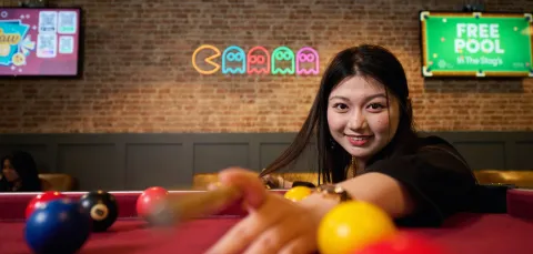 A student playing pool in a students' union bar. The student has long dark hair, and the background shows a brick wall with televisions and a neon sign in the shape of characters from the game Pac-Man.