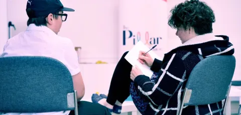 Two young adults sitting with their backs to the camera. One has their feet up on a table and is writing on a note pad resting on their knee. The other has a baseball cap on and is watching the person writing. there is a Pillar logo on the wall.