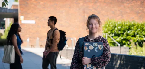 A student smiling while stood outside and holding a closed laptop. There are two more students talking to each other in the background.